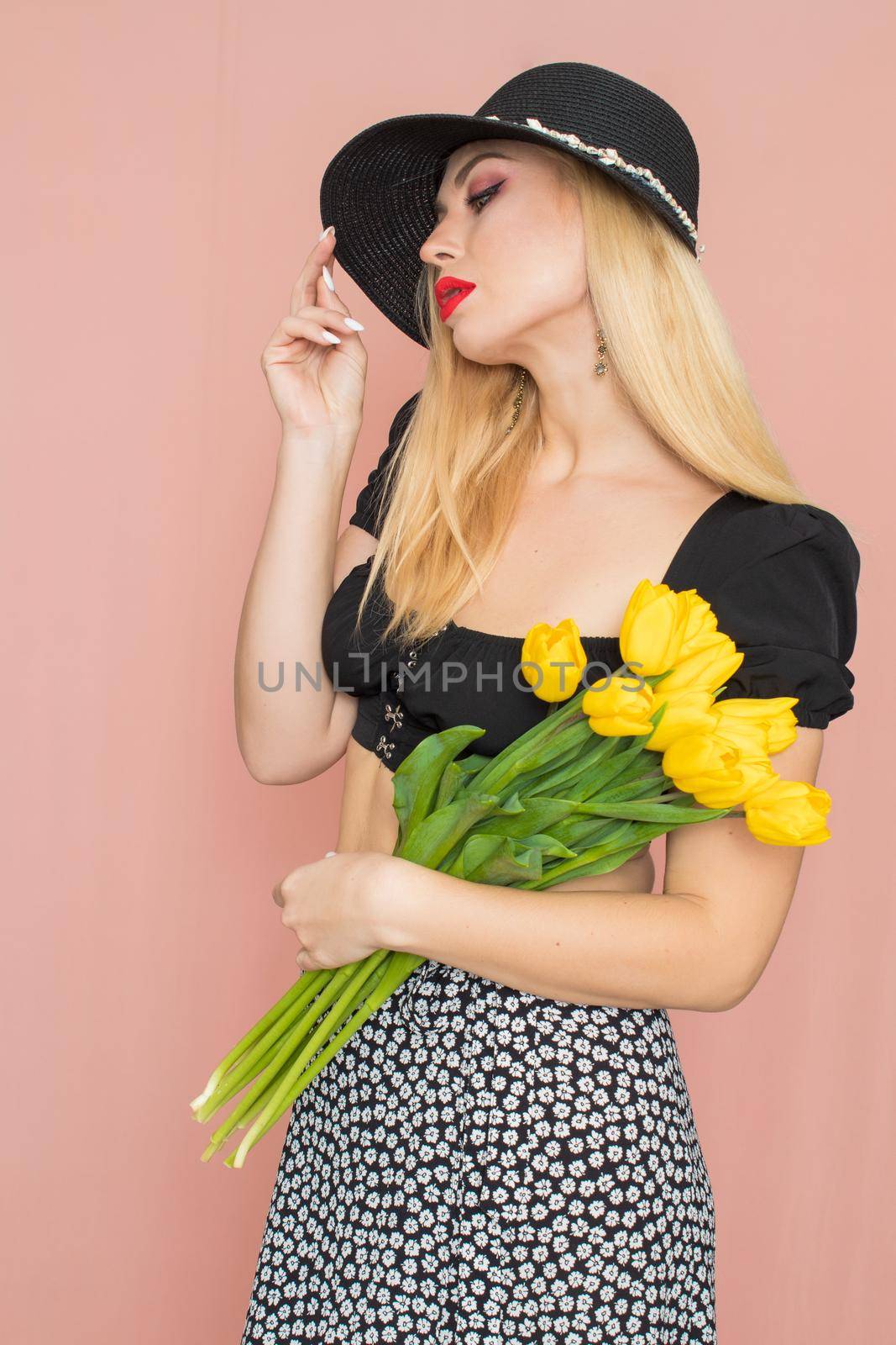 Summer fashion portrait blonde woman. Sexy look in black top and skirt, wearing hat. Red lips. Holding yellow tulips in her hands