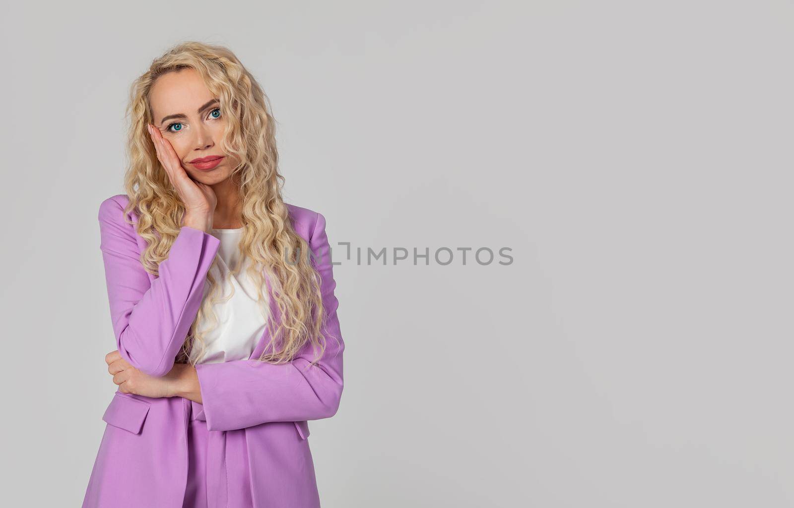 You spend my time stupid talking. Tired and bored, a cute blonde woman with blue eyes, leaning against her palm, standing over a gray wall with an indifferent annoyed expression. looks at the camera.