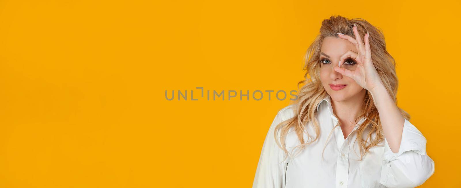 Blond woman feeling optimistic happy showing ok circle over eye smiling happily. feels happy optimistic showing ok, good gesture okay with an eye sign smiling happily standing yellow background
