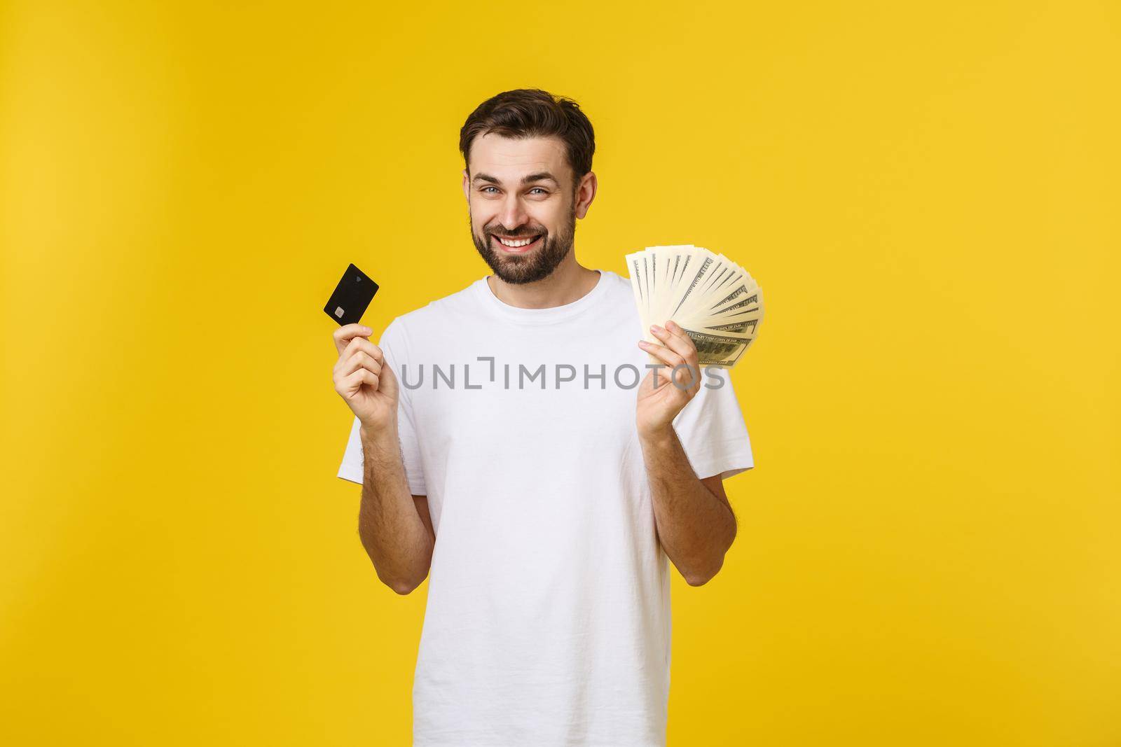 Young handsome happy smiling man holding banking card and cash in his hands isolated on yellow background. by Benzoix