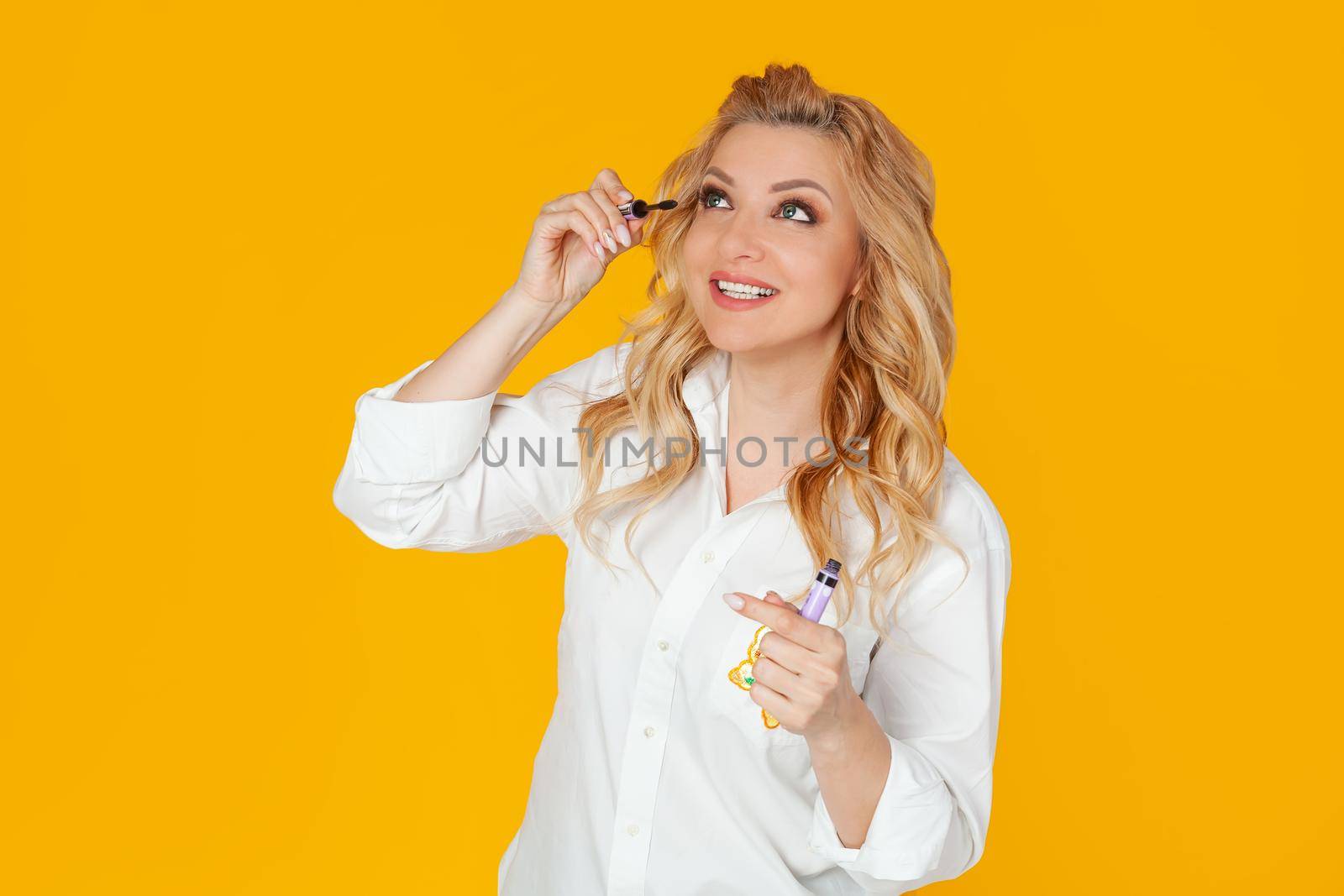 Blonde in white shirt, middle aged European woman smiles broadly, holding and applying mascara, standing against yellow background