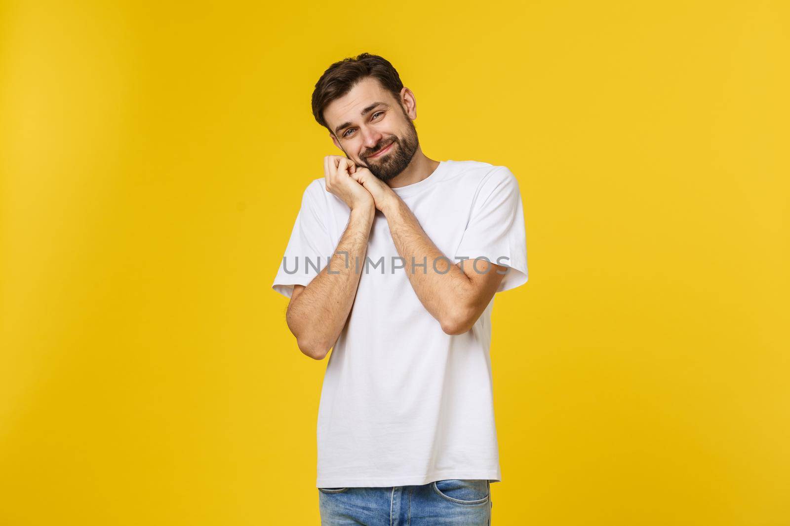 Young man looks to the camera and smiles shy. He's got his hands on cheek. Isolated, with a beard and a white T-shirt.