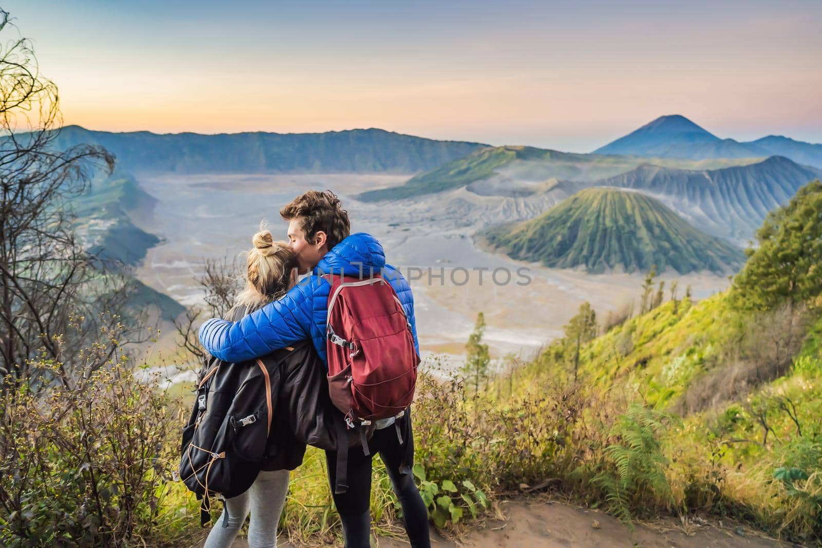 Young couple man and woman meet the sunrise at the Bromo Tengger Semeru National Park on the Java Island, Indonesia. They enjoy magnificent view on the Bromo or Gunung Bromo on Indonesian, Semeru and other volcanoes located inside of the Sea of Sand within the Tengger Caldera. One of the most famous volcanic objects in the world. Travel to Indonesia concept by galitskaya