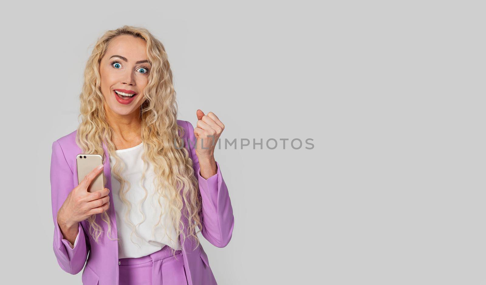 A happy triumphant woman holds a smartphone, fist, and an enthusiastic smile receives great fantastic news using a text message on her mobile phone. Wins the online lottery, receives the first prize.