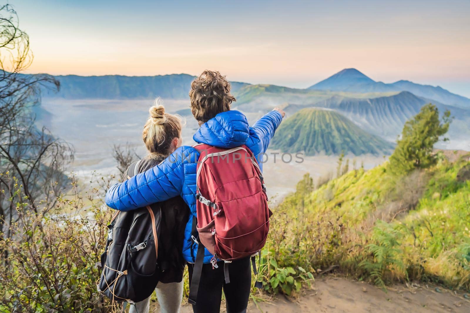 Young couple man and woman meet the sunrise at the Bromo Tengger Semeru National Park on the Java Island, Indonesia. They enjoy magnificent view on the Bromo or Gunung Bromo on Indonesian, Semeru and other volcanoes located inside of the Sea of Sand within the Tengger Caldera. One of the most famous volcanic objects in the world. Travel to Indonesia concept by galitskaya