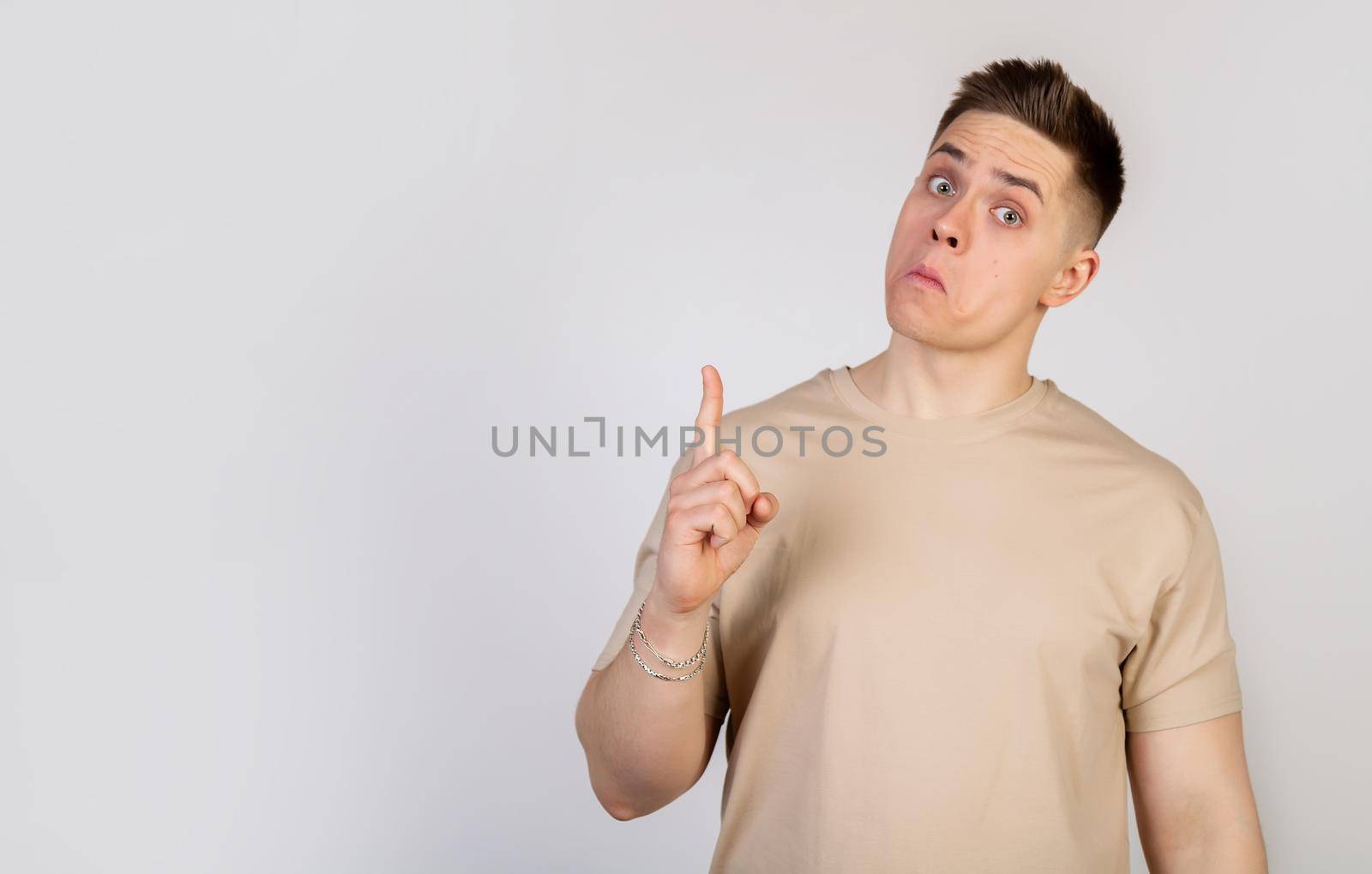 A handsome guy with a beautiful haircut looks at the camera with a funny facial expression, holding his lips and pointing his finger up. Copy space
