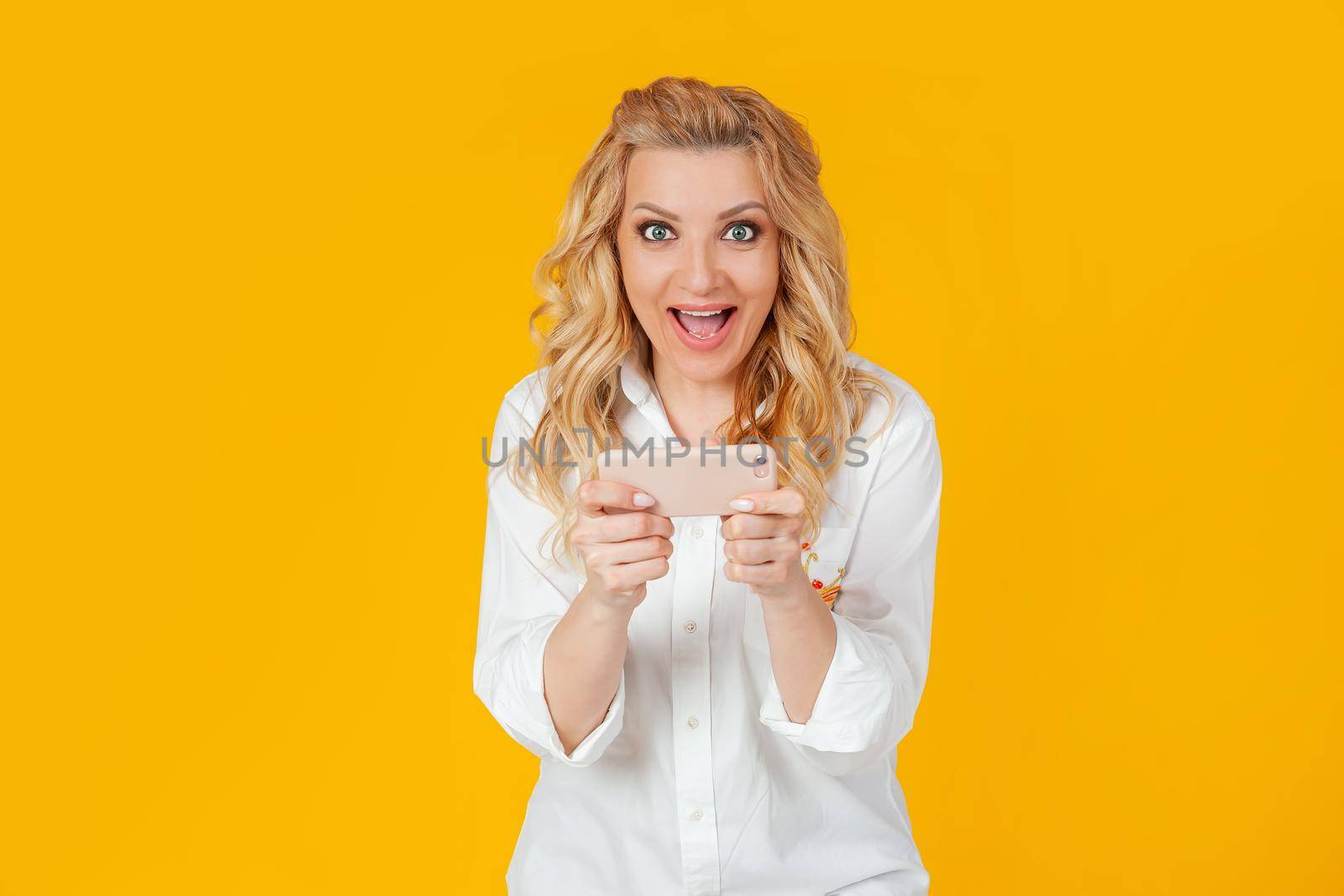 Excited European blonde woman in white shirt, looks surprised and excited, plays awesome new smartphone game, screams and smiles happy, wins. Standing on a yellow background
