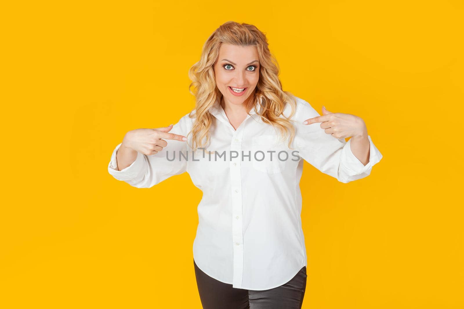Amazed speechless impressed happy European blonde woman in white shirt pointing at herself shocked, with open mouth, drooping jaw, amazed, to receive an unexpected great offer on a yellow background.