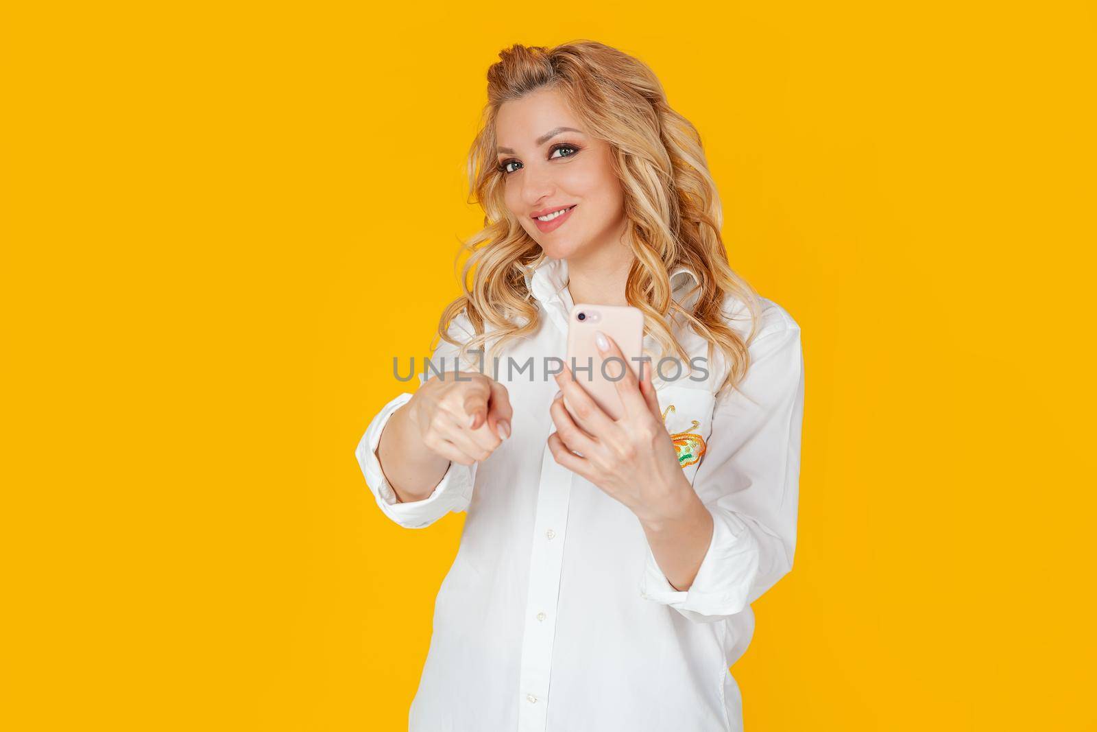 Portrait of charismatic blonde European woman in white shirt taking photo or recording video with you, pointing at camera, holding smartphone and smiling cheerfully. Standing on a yellow background