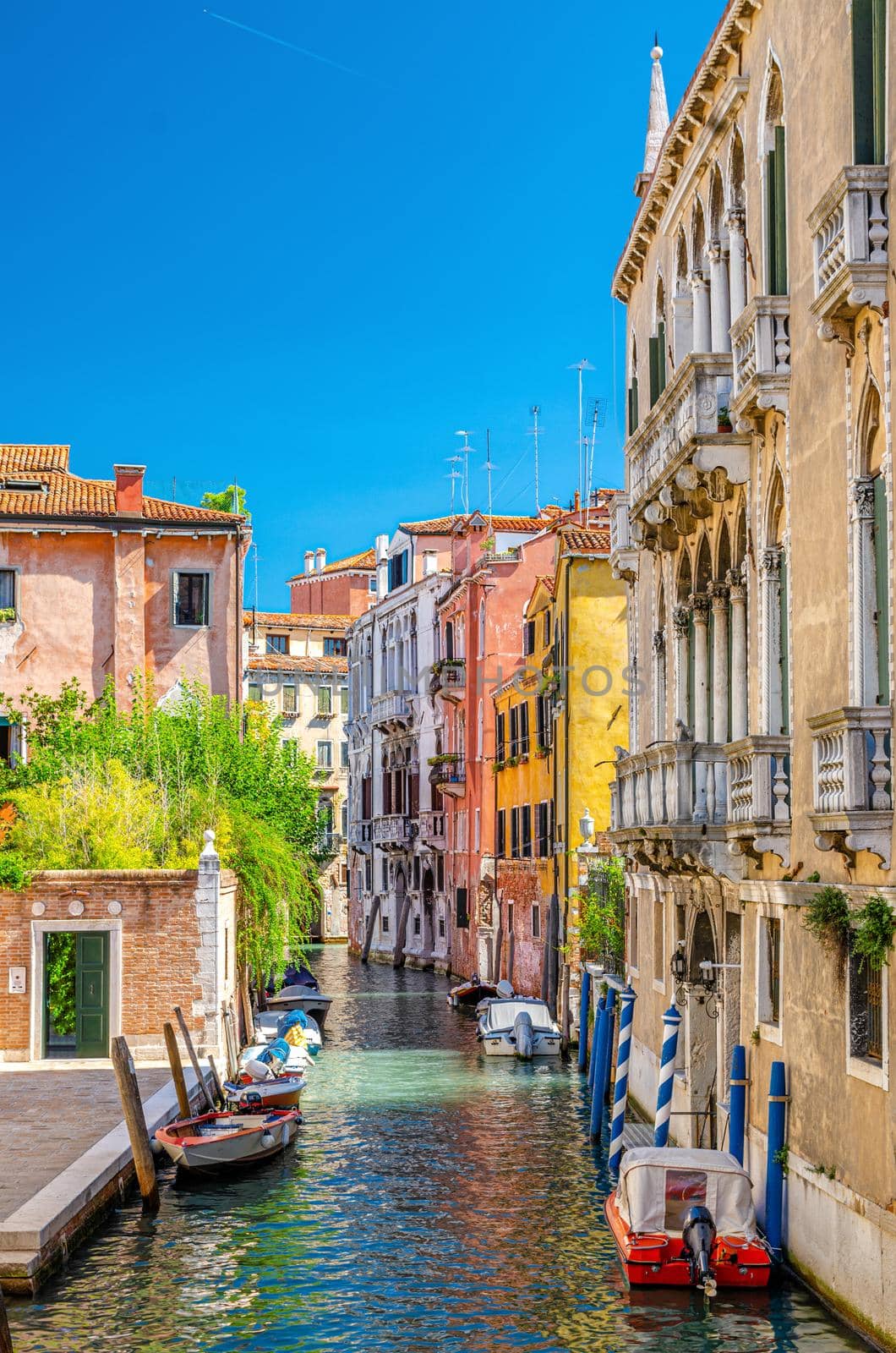 Typical Venice narrow water canal with moored boats between old colorful buildings with balconies and brick walls, Veneto Region, Northern Italy. Traditional Venetian cityscape, blue sky background.