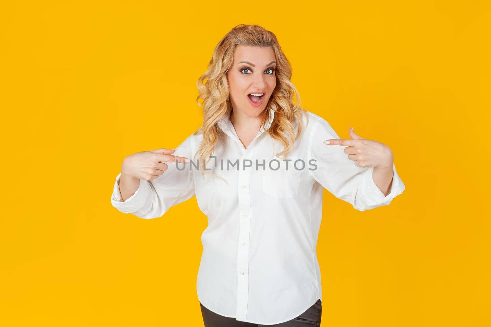 Amazed speechless impressed happy European blonde woman in white shirt pointing at herself shocked, with open mouth, drooping jaw, amazed, to receive an unexpected great offer on a yellow background.