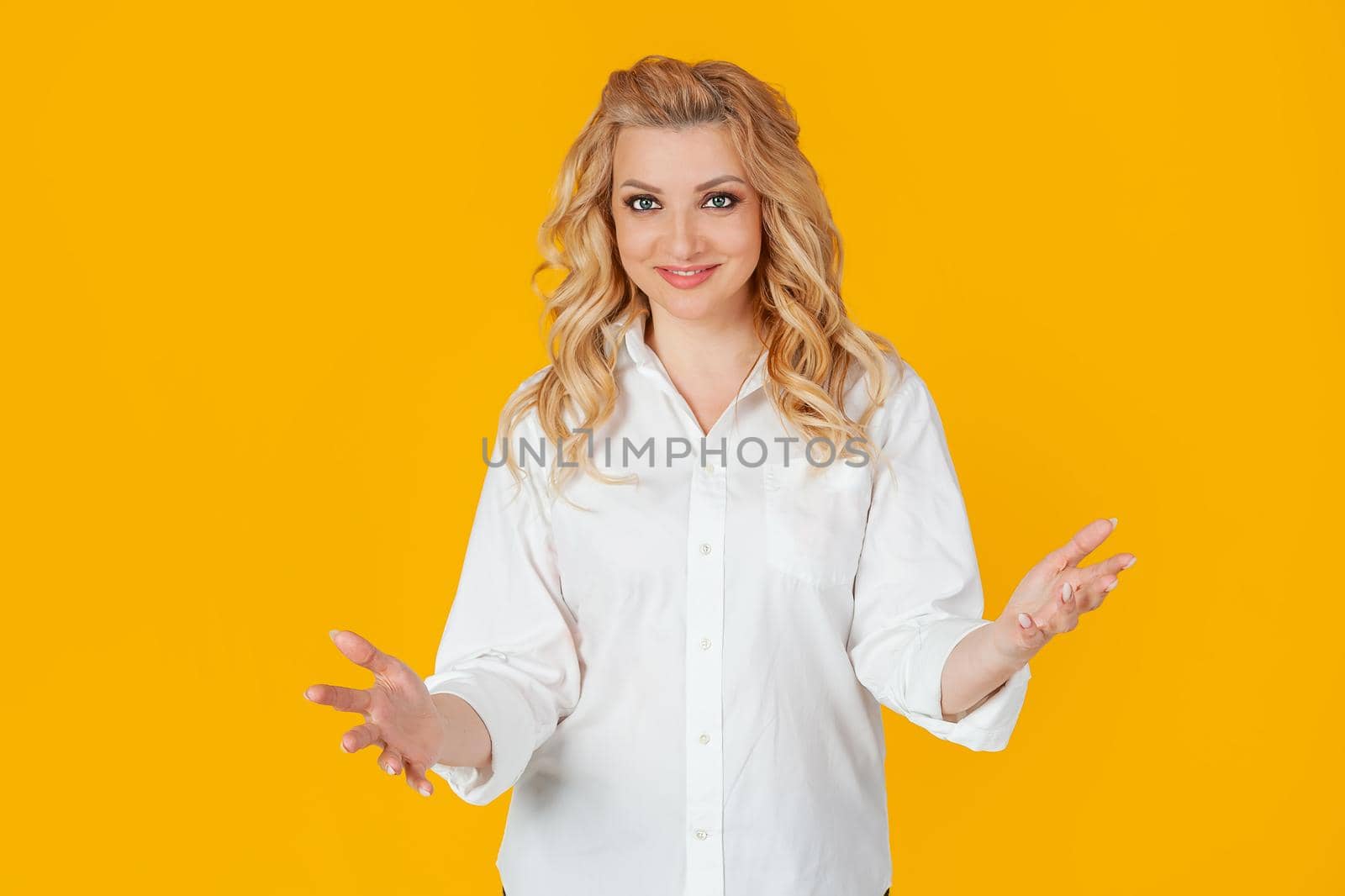 Sociable cute woman in white shirt, middle aged European girl, extends her arms to the camera in greeting and warm welcome, wanting to hug or hug, standing against yellow background.
