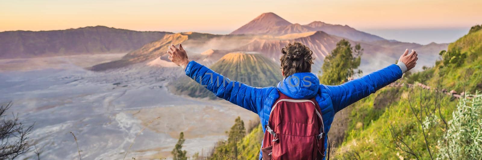 BANNER, LONG FORMAT Young man meets the sunrise at the Bromo Tengger Semeru National Park on the Java Island, Indonesia. He enjoys magnificent view on the Bromo or Gunung Bromo on Indonesian, Semeru and other volcanoes located inside of the Sea of Sand within the Tengger Caldera. One of the most famous volcanic objects in the world. Travel to Indonesia concept.