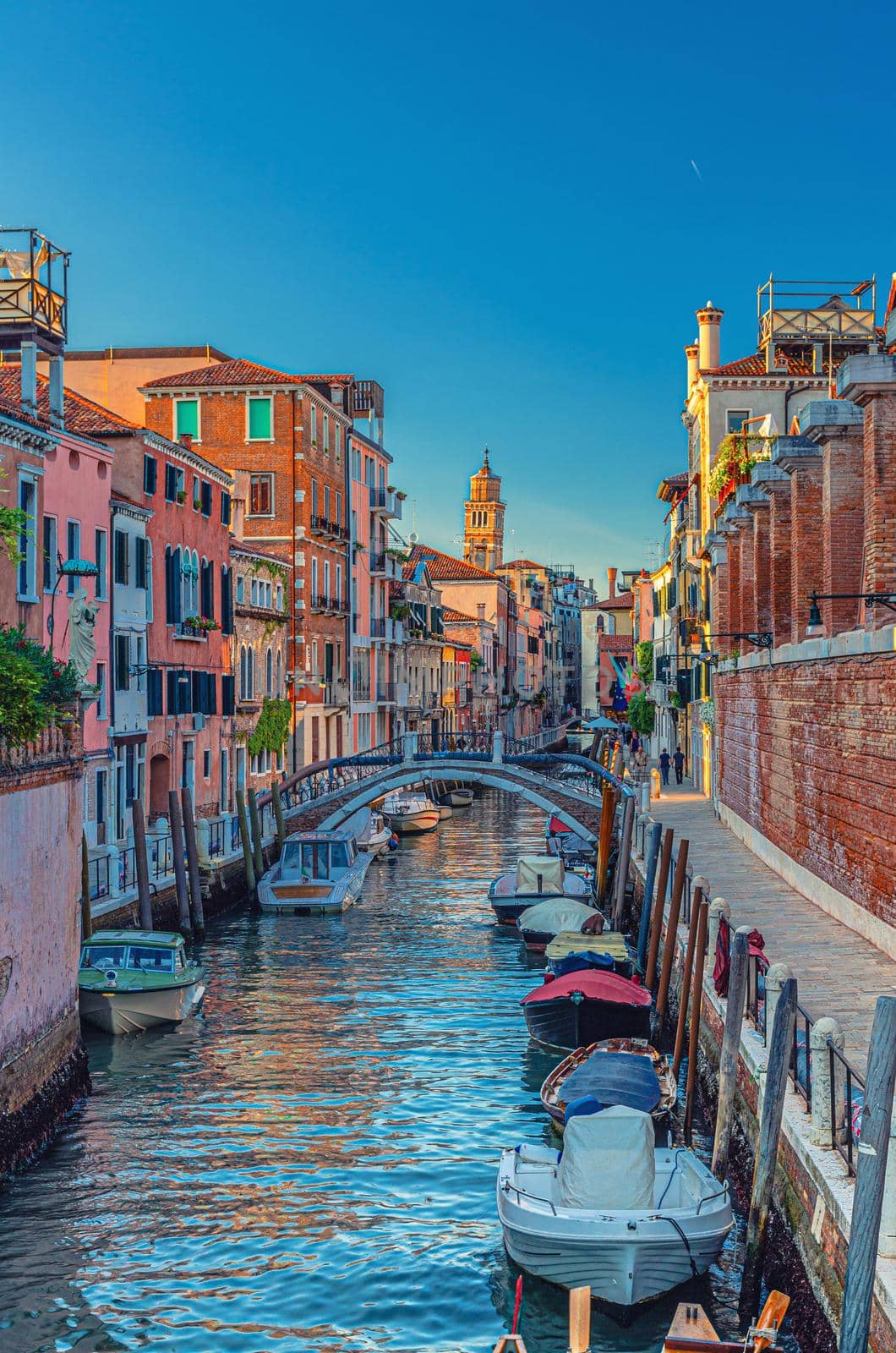 Venice cityscape with narrow water canal and colorful boats moored near fondamenta embankment, Veneto Region, Northern Italy. Typical Venetian view at sunset, vertical view, blue sky background