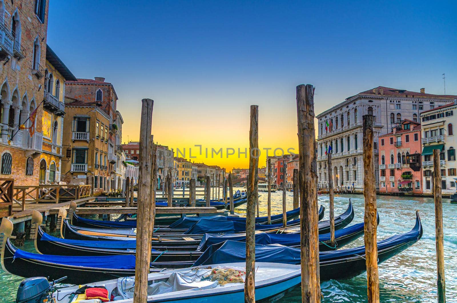 Gondolas moored docked on pier of Grand Canal waterway in Venice. Wooden poles and baroque style buildings along Canal Grande. Amazing Venice cityscape at sunset twilight. Veneto Region, Italy