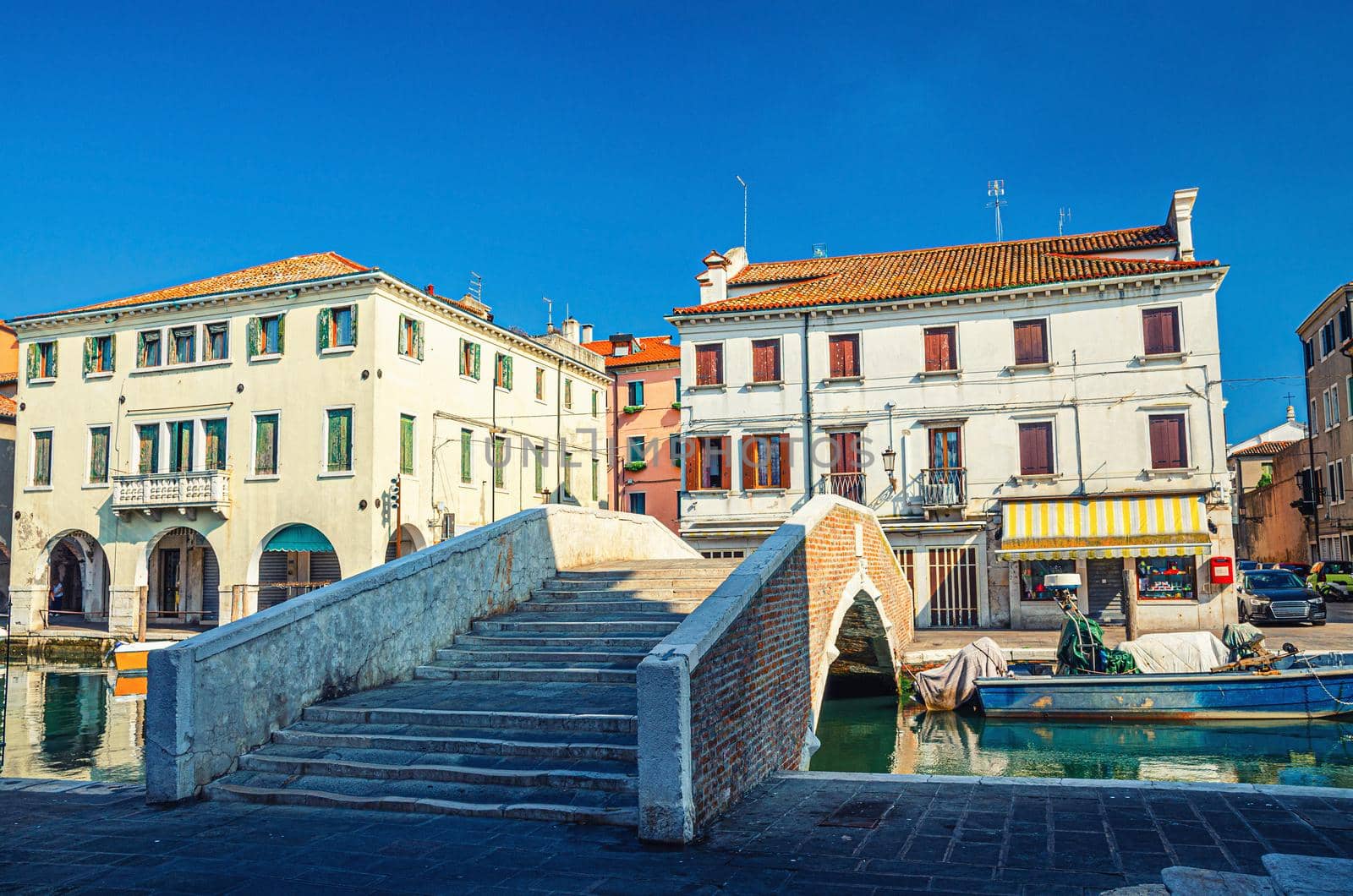 Stone brick bridge Ponte Pescheria across Vena water canal and old buildings in historical centre of Chioggia town, blue sky background in summer day, Veneto Region, Northern Italy