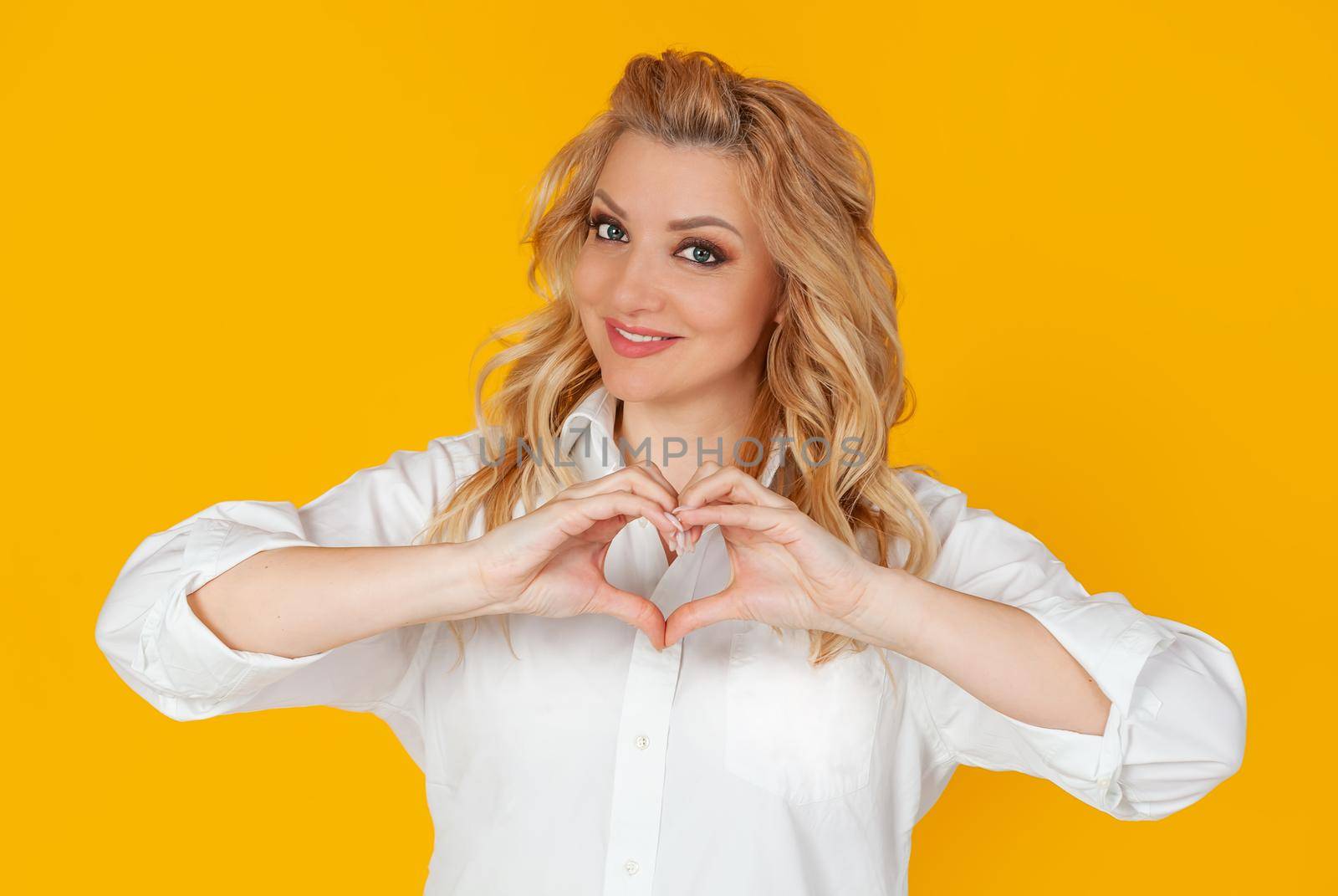 Blonde in white shirt, middle aged European girl showing heart gesture acknowledging sympathy, smiling friendly at camera, on yellow background by ViShark