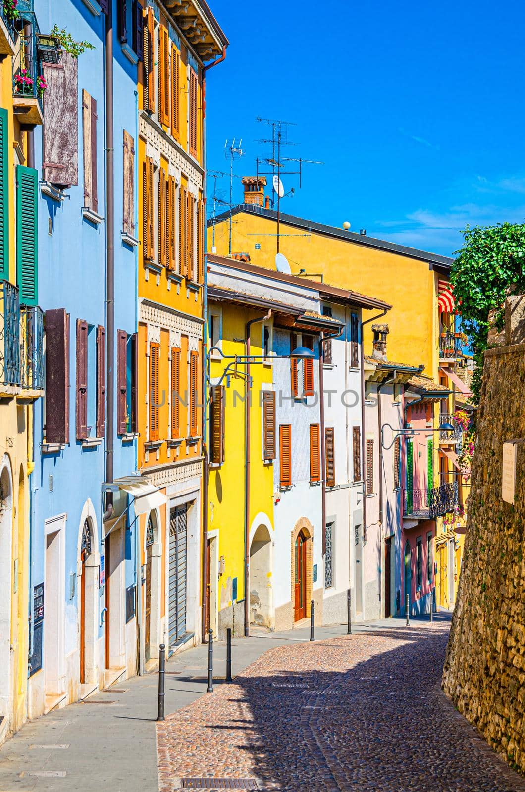 Typical narrow street in old historical centre of Desenzano del Garda town with cobblestone road and traditional colorful buildings with shutter windows, vertical view, Lombardy, Northern Italy