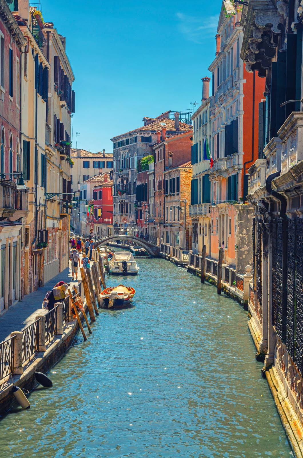 Venice cityscape with narrow water canal with moored boats between old colorful multicolored buildings and stone promenade, Veneto Region, Northern Italy. vertical view, blue sky background.
