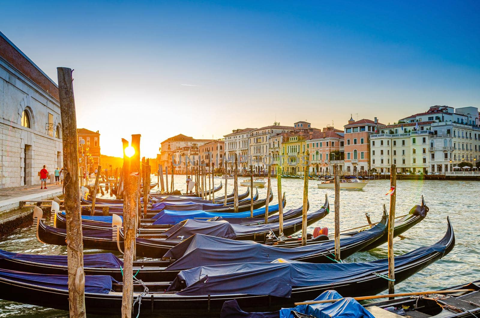 Gondolas moored docked on pier of Grand Canal waterway in Venice. Baroque style buildings along Canal Grande background. View against sun. Amazing Venice cityscape at sunset. Veneto Region, Italy