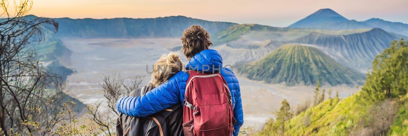 BANNER, LONG FORMAT Young couple man and woman meet the sunrise at the Bromo Tengger Semeru National Park on the Java Island, Indonesia. They enjoy magnificent view on the Bromo or Gunung Bromo on Indonesian, Semeru and other volcanoes located inside of the Sea of Sand within the Tengger Caldera. One of the most famous volcanic objects in the world. Travel to Indonesia concept.