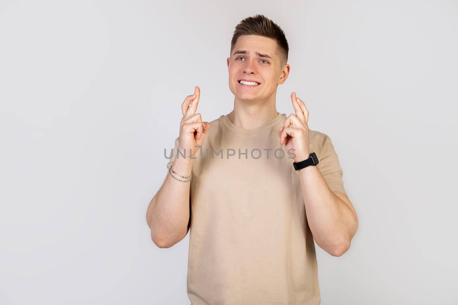 A handsome young guy crosses his fingers with a worried expression, asking God please, keep your fingers crossed with a pitiful look, hoping that the boss did not notice that he is late for work.