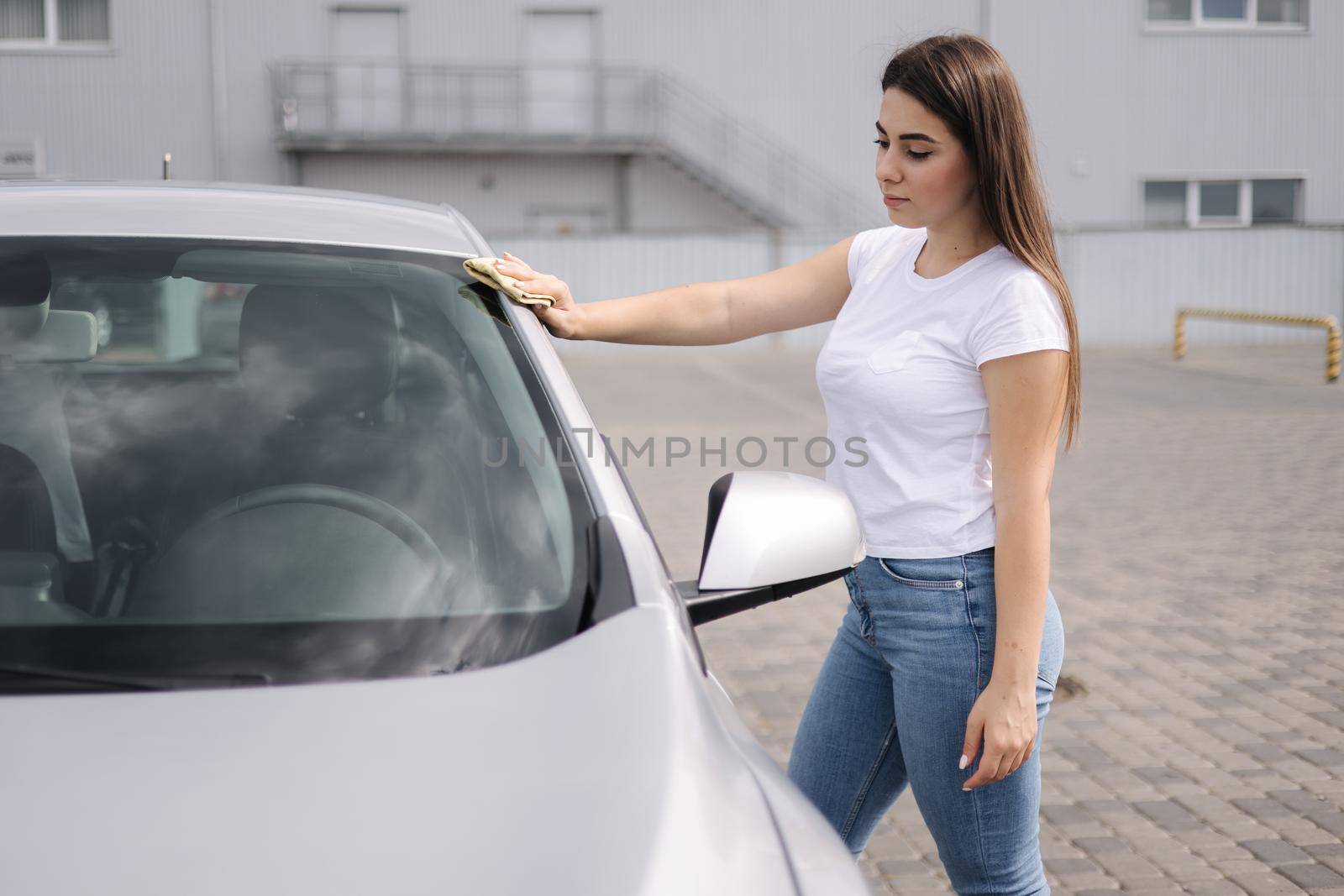 Front view of attractive happy joyful female driver washing her car's headlights with special rag in self-service car wash.