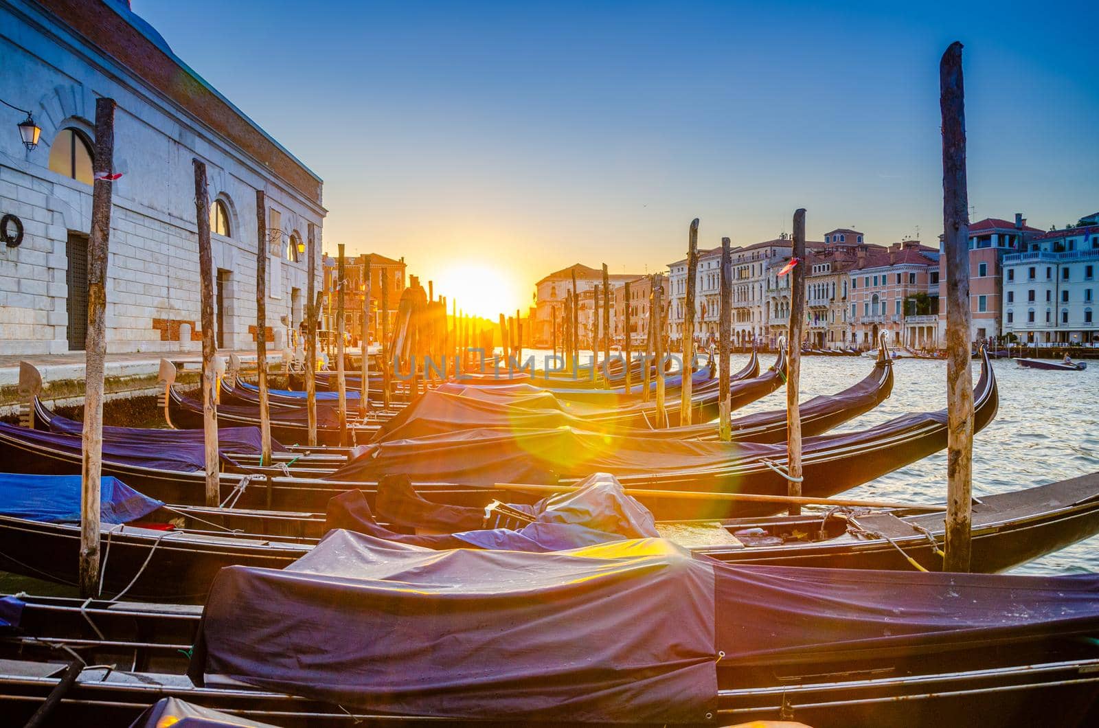 Gondolas moored docked on pier of Grand Canal waterway in Venice. Baroque style buildings along Canal Grande background. View against sun. Amazing Venice cityscape at sunset. Veneto Region, Italy