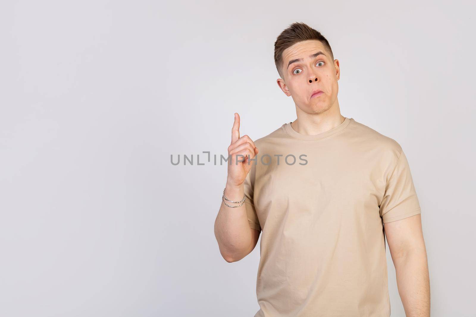 A handsome guy with a beautiful haircut looks at the camera with a funny facial expression, holding his lips and pointing his finger up. Copy space