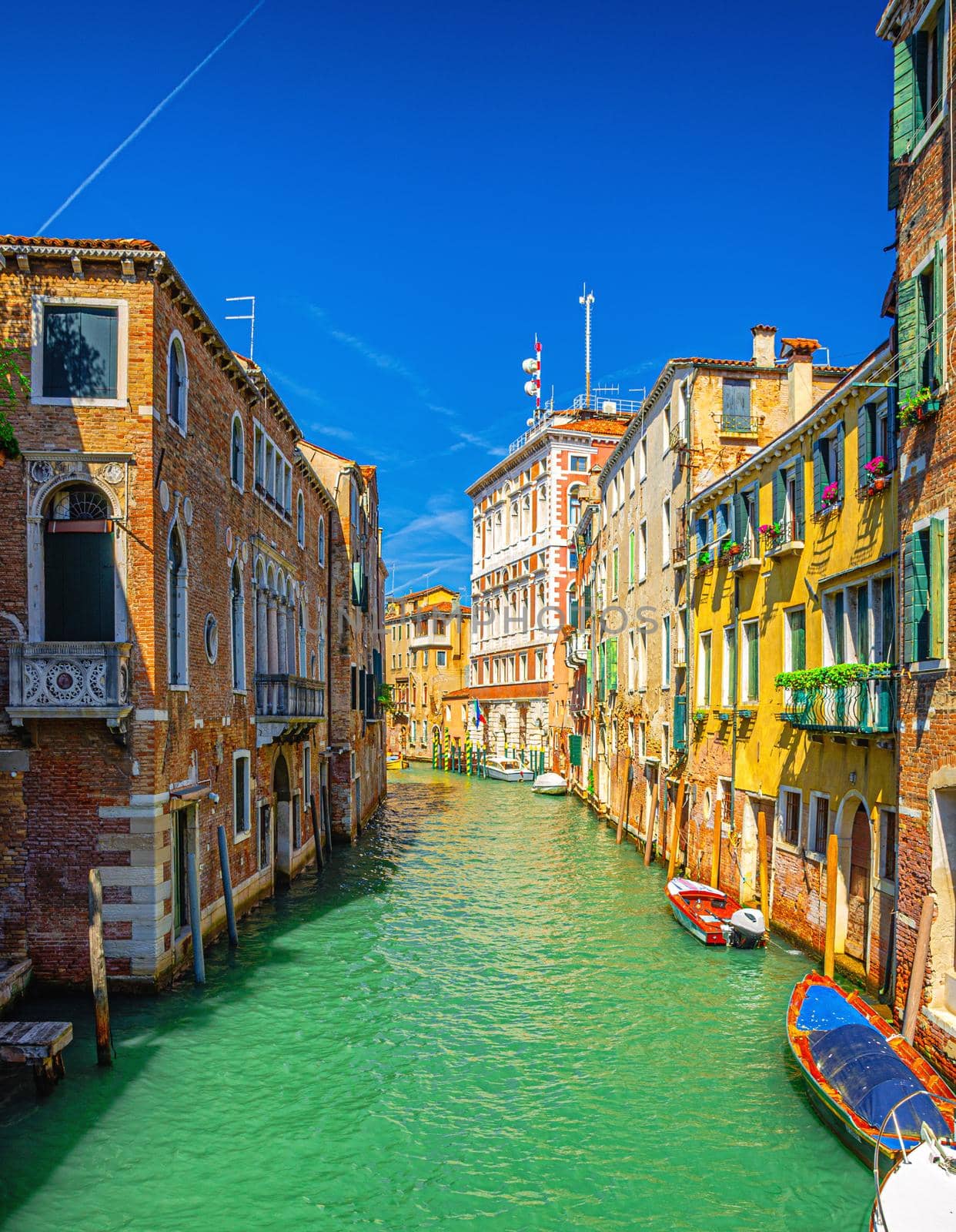 Venice cityscape with narrow water canal with colorful boats moored near old multicolored buildings, Veneto Region, Northern Italy. Typical Venetian view, vertical view, blue sky background