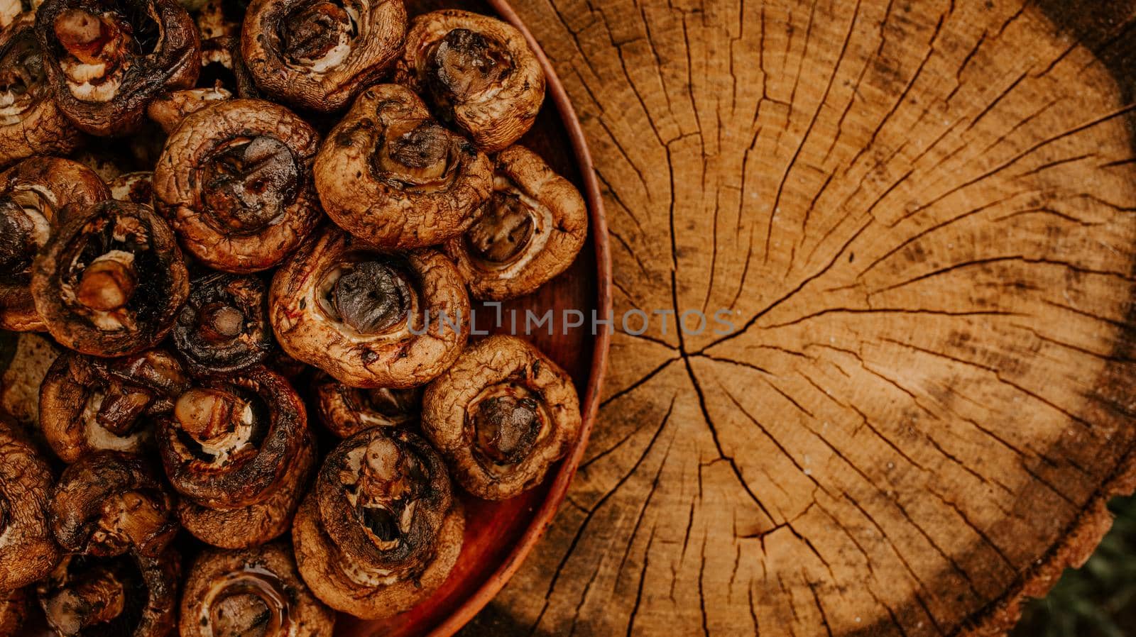 Cooked baked champignon mushrooms lie on a brown clay plate on a cracked brown hemp. Summer green blurred grass background.