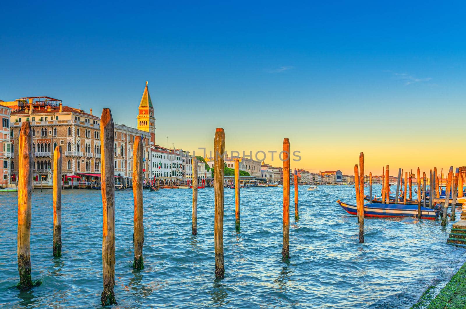 Venice evening view with wooden poles and boats near pier of Grand Canal waterway at sunset, Campanile bell tower and row of baroque style buildings in San Marco sestiere, Veneto region, Italy