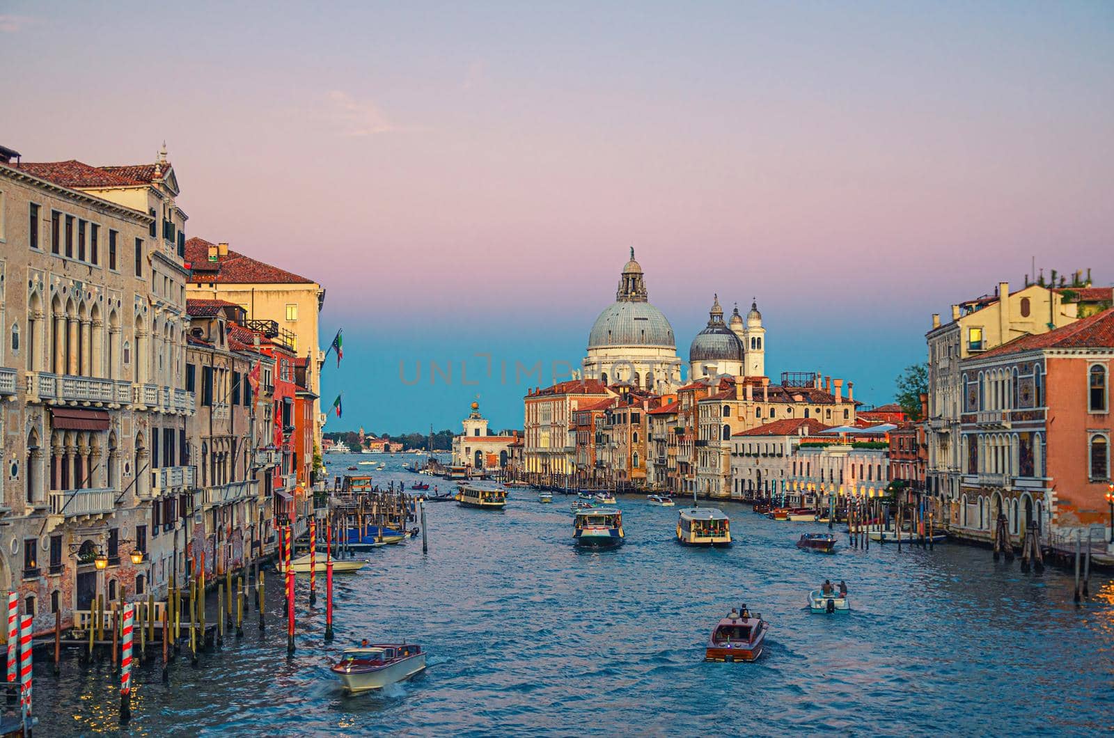 Venice cityscape with Grand Canal waterway. Buildings with evening lights along Grand Canal. Santa Maria della Salute Roman Catholic church on Punta della Dogana at twilight. Veneto Region, Italy.