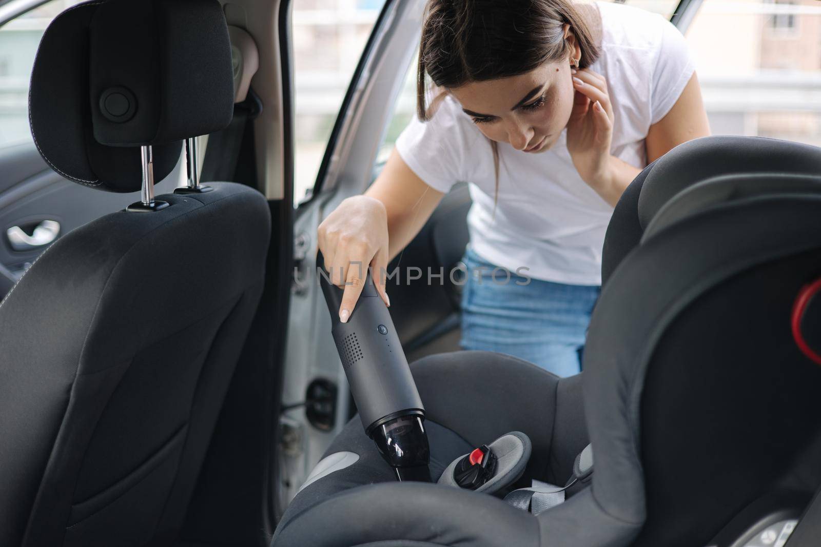 Close-up of female using portable vacuum cleaner in her car. Baby ca seat cleaning. Woman vacuuming seats. Dust and dirt removal.