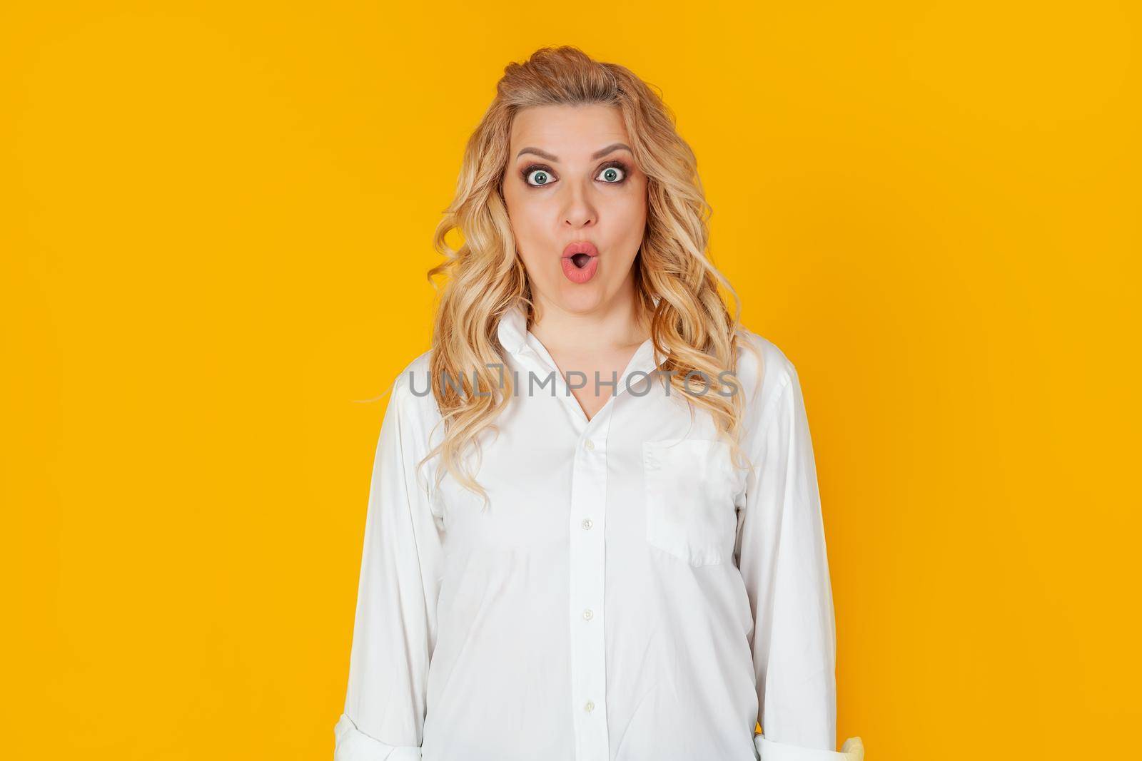 An excited impressed woman, a blonde in a white shirt, with a surprised open mouth, looks frightened at the camera, expressing amazement from the shock. Isolated on a yellow background.