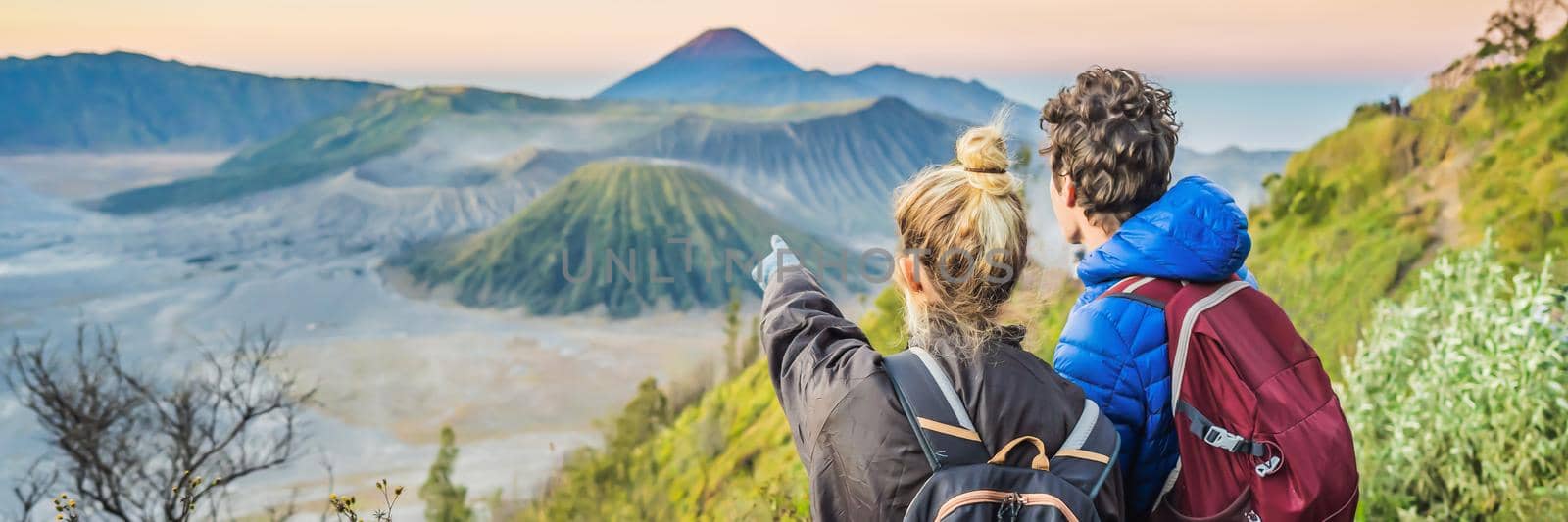 BANNER, LONG FORMAT Young couple man and woman meet the sunrise at the Bromo Tengger Semeru National Park on the Java Island, Indonesia. They enjoy magnificent view on the Bromo or Gunung Bromo on Indonesian, Semeru and other volcanoes located inside of the Sea of Sand within the Tengger Caldera. One of the most famous volcanic objects in the world. Travel to Indonesia concept by galitskaya