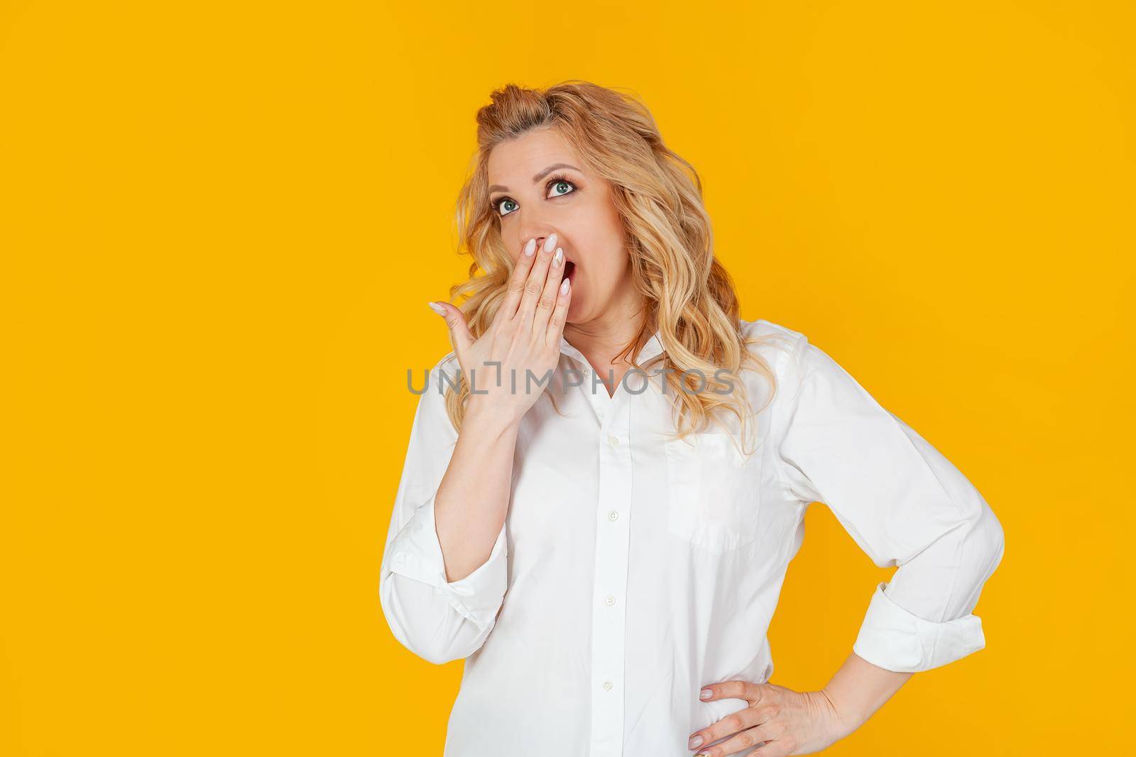 Portrait of a woman in a white shirt, a middle-aged European girl, yawning and covering her open mouth, with a boring face, wakes up early, stands on a yellow background.