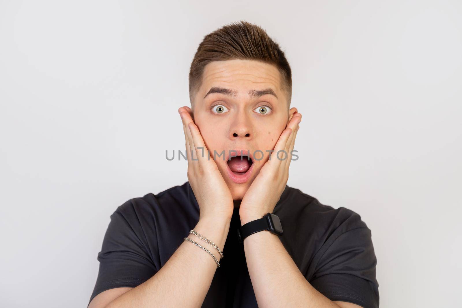 Shocked young man holding his hands on his cheeks, looking with surprised eyes and opened his mouth to the camera. Guy marveling at open mouth, gaze with astonishment, jaw drop from news.