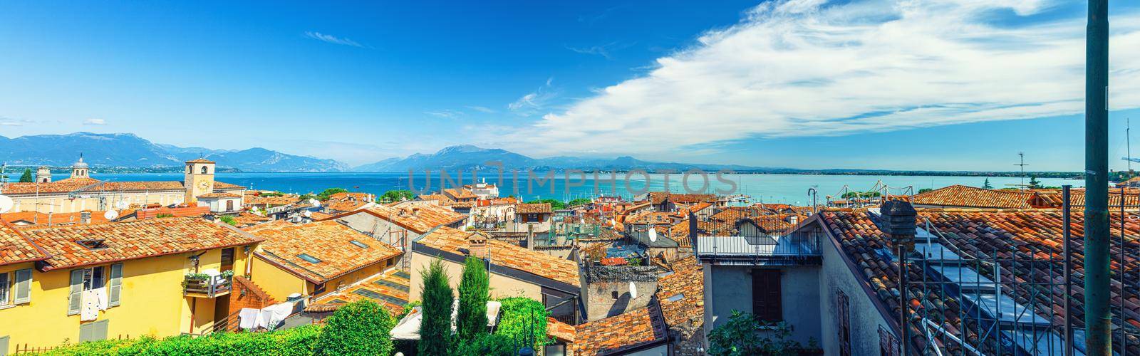 Panorama of Desenzano del Garda town with red tiled roof buildings, Garda Lake water, Monte Baldo mountain range, Sirmione peninsula, Lombardy, Northern Italy. Aerial panoramic view of Desenzano