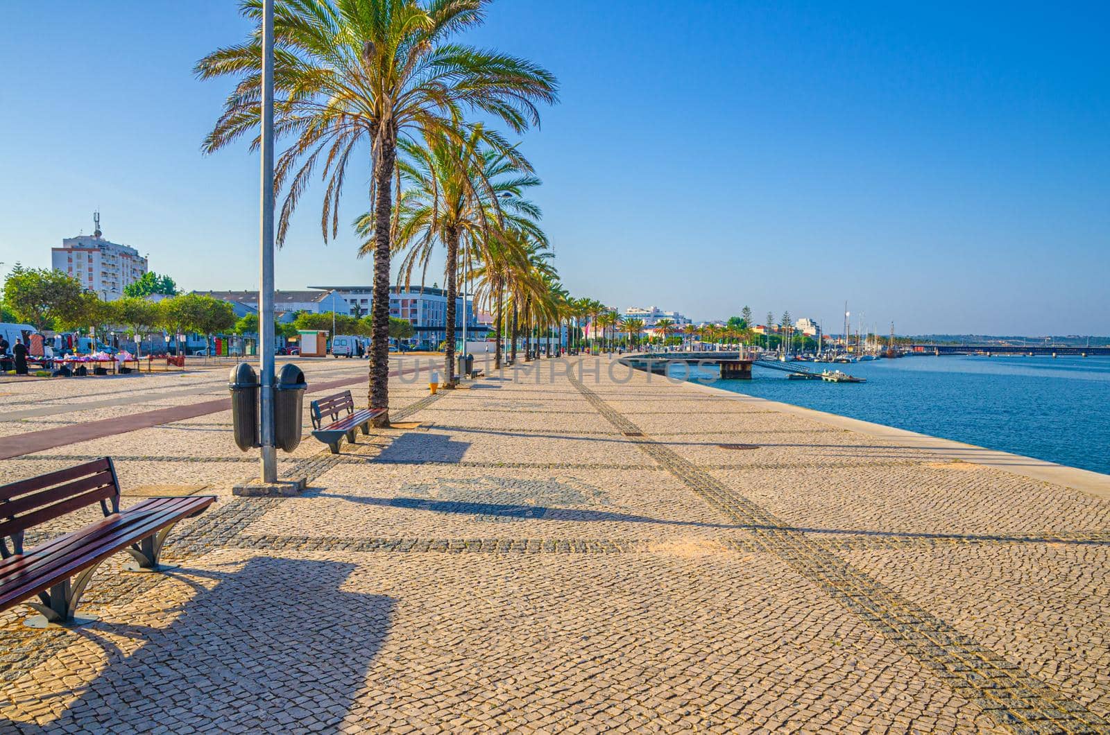 Ribeirinha de Portimao town, cobblestone embankment promenade of Arade River in city centre with palm trees and benches, blue sky background, Faro district, Algarve region, Portugal