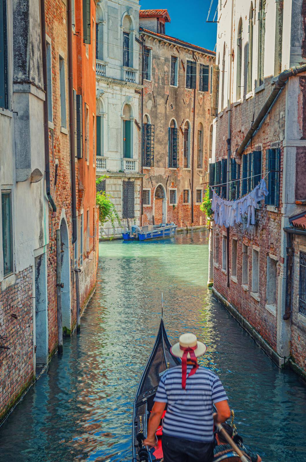 Gondola sailing narrow canal in Venice between old buildings with brick walls. Gondolier dressed traditional white and blue striped short-sleeved polo shirt and boater straw hat with red ribbon.