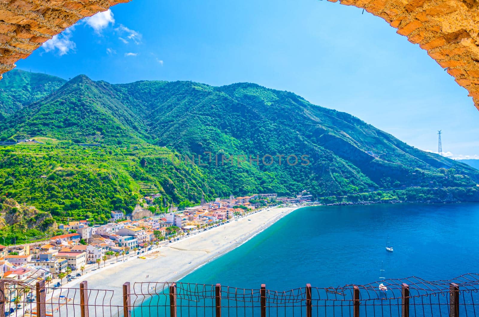 Cityscape of Scilla village with sandy beach of Tyrrhenian sea bay gulf coast shore, aerial view through stone arch of beautiful seaside town with green hill background, Calabria, Southern Italy