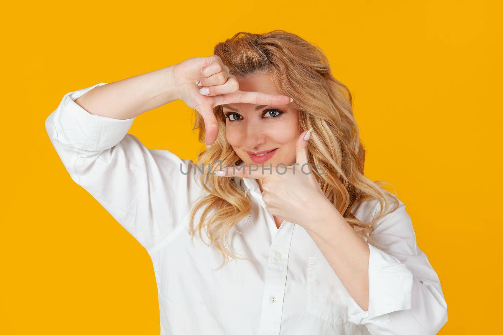 A blonde in a white shirt, middle aged European woman, a designer creating a new project, looks at the footage and smiles, and stands against a yellow background to create the perfect shot