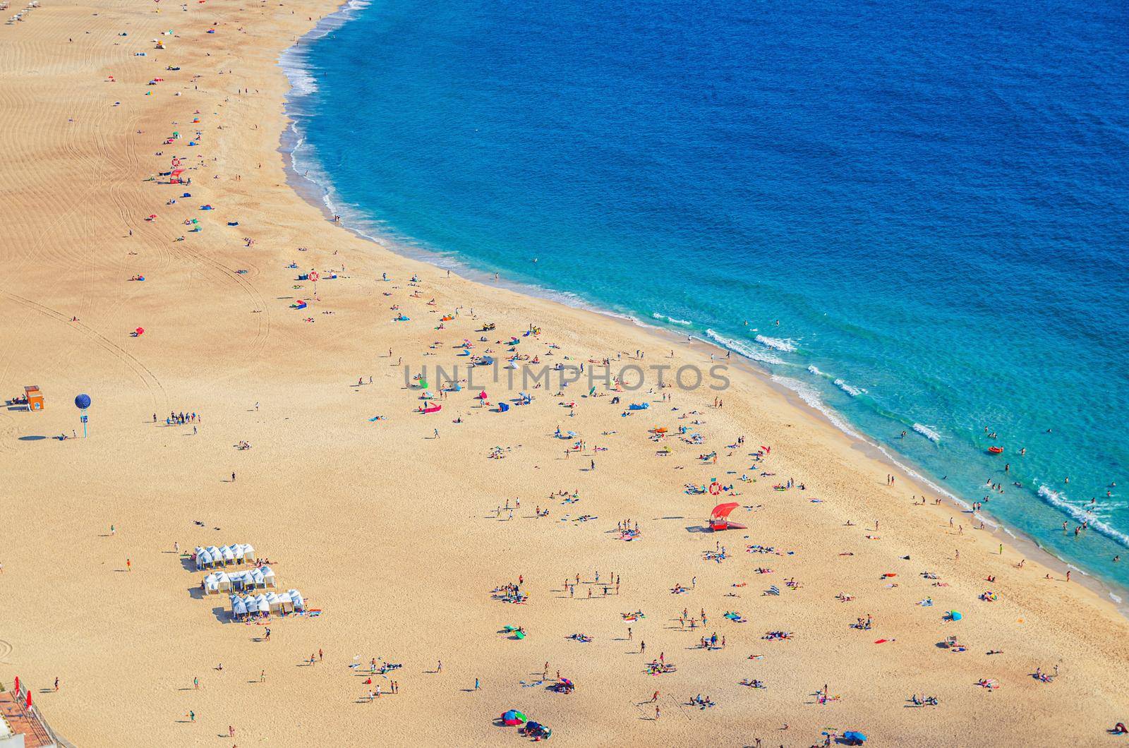 Top aerial view of sandy beach with people tourists sunbathing and Atlantic Ocean azure blue water, Praia da Nazare town coastline with waves, Leiria District, Oeste region, Portugal