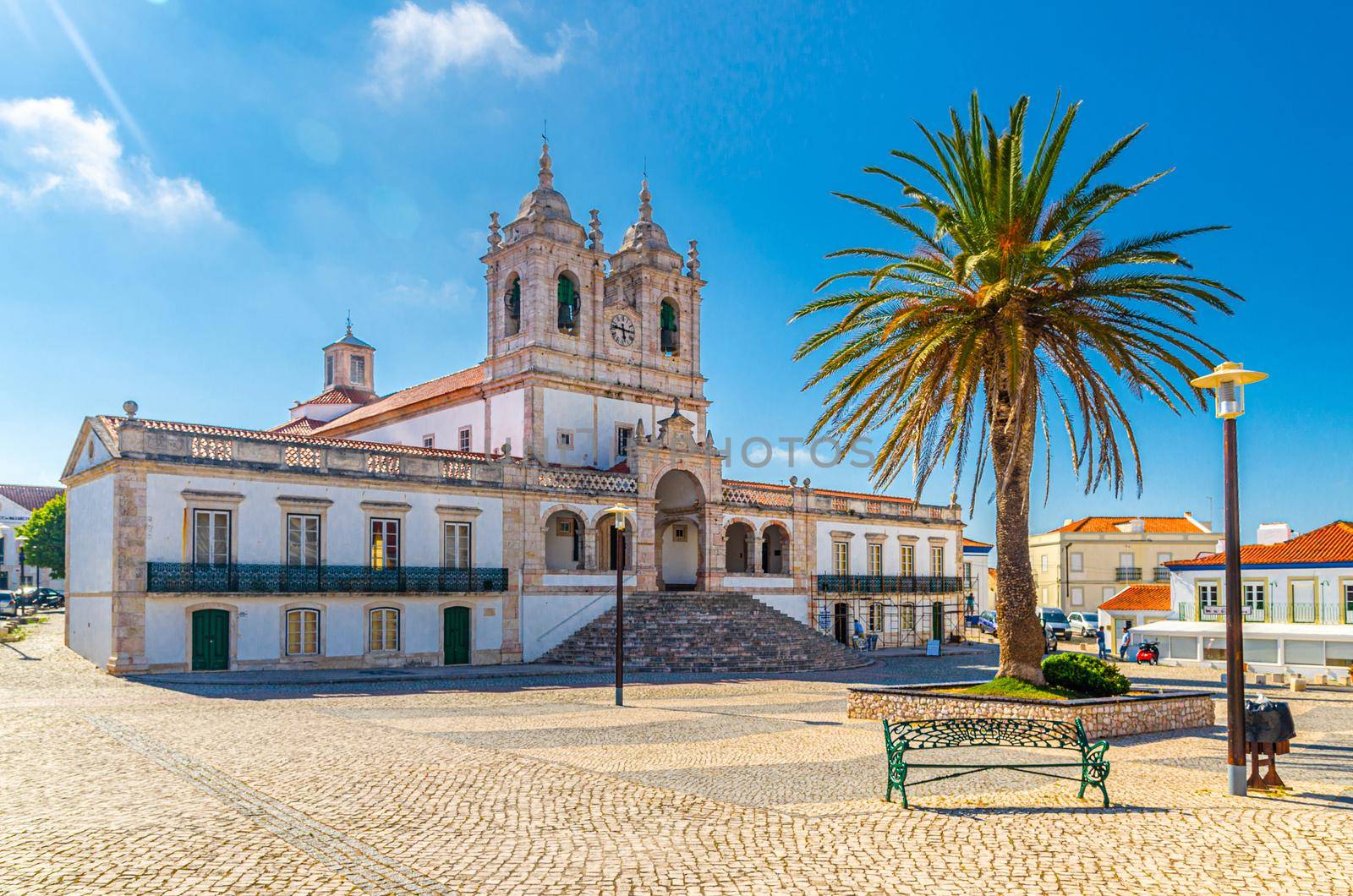 Sanctuary of Our Lady of Nazare catholic church in cobblestone square with palm trees in Sitio hilltop da Nazare town, clear blue sky in sunny summer day, Leiria District, Oeste region, Portugal