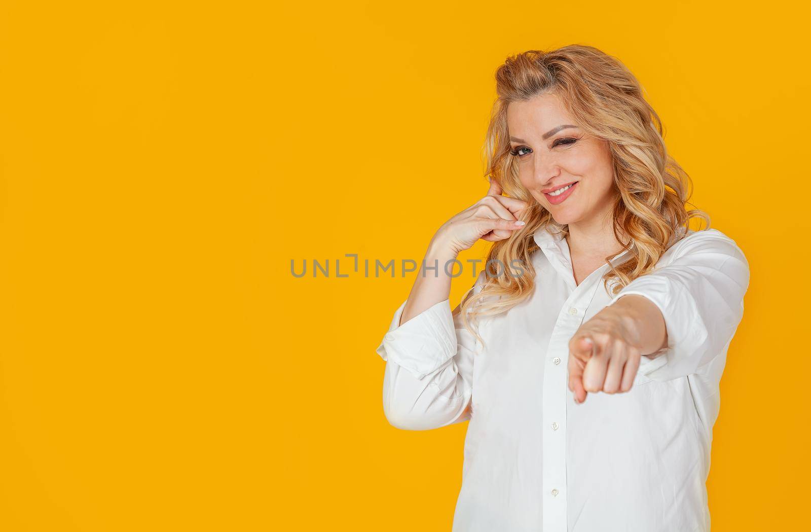 Portrait of a beautiful blonde woman in a white shirt, pointing to the camera, smiling and making a phone gesture, as if asking for a number, flirting with someone, standing on a yellow background.