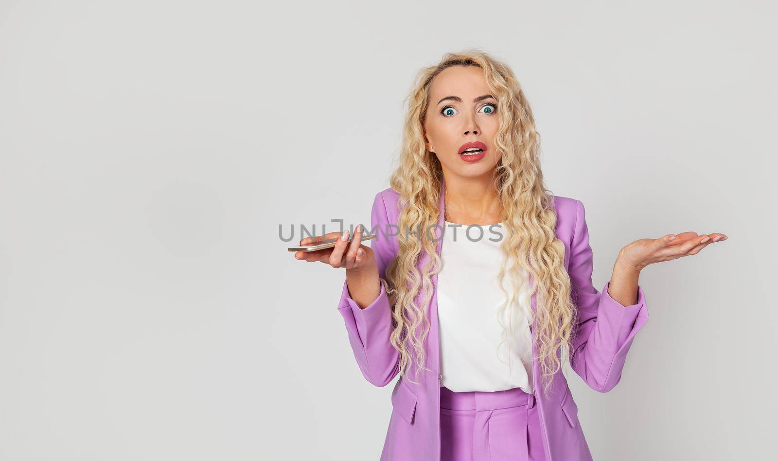 An embarrassed and excited blonde woman does not know what she did wrong, shrugs and shrugs her hands upset, holds a smartphone, explains something to her friend, looks nervous