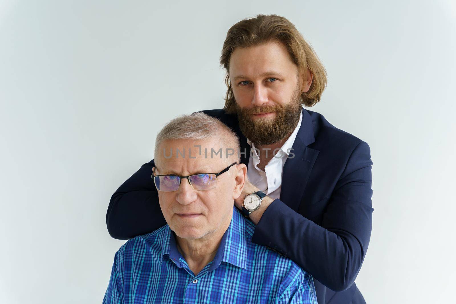 the son in a stylish suit leans on the shoulders of his father, Father and son in white background, both men look into the camera, an elderly man in glasses, young man with a beard and long hair by vladimirdrozdin