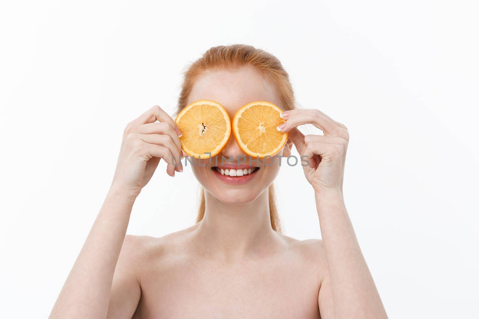 Portrait of a cheerful young girl holding two slices of an orange at her face over white wall background.