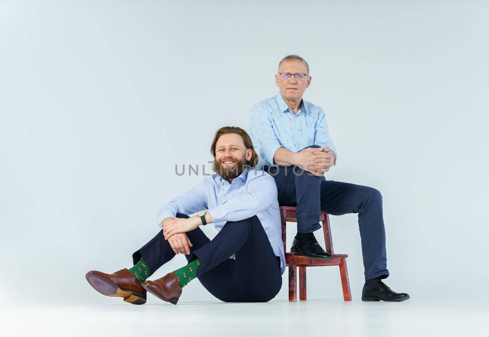 Father sits on the chair and son sits on white background, both men look into the camera, an elderly man in glasses with diopters, a stylish young man with a beard and long hair. High quality photo